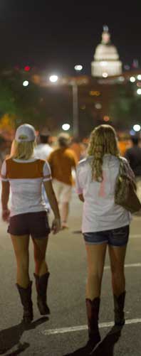 Two women walk behind a crowd on the street at night; capitol dome in background